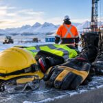 Industrial worker wearing cold-weather PPE at a snowy Alaska worksite with hard hats, safety goggles, insulated gloves, hearing protection, and work boots.